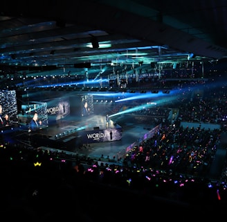 A large indoor arena filled with an audience watching a live performance. The stage features large screens displaying a performer and graphics, with bright spotlights and ambient lighting creating a dynamic atmosphere. The crowd is holding glow sticks, adding vibrant colors to the scene.