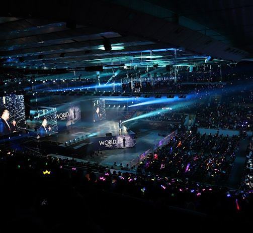 A large indoor arena filled with an audience watching a live performance. The stage features large screens displaying a performer and graphics, with bright spotlights and ambient lighting creating a dynamic atmosphere. The crowd is holding glow sticks, adding vibrant colors to the scene.