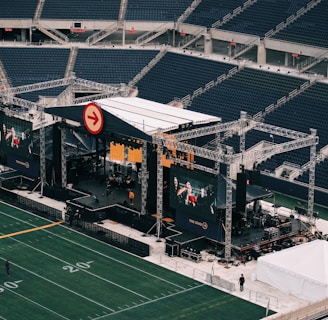 A large outdoor stage is set up inside a stadium, equipped with tall scaffolding, lights, and multiple large display screens. The seats in the stadium are mostly empty. The field is marked with numbers and lines typical of a sports venue. A few people are visible near the stage and on the field.