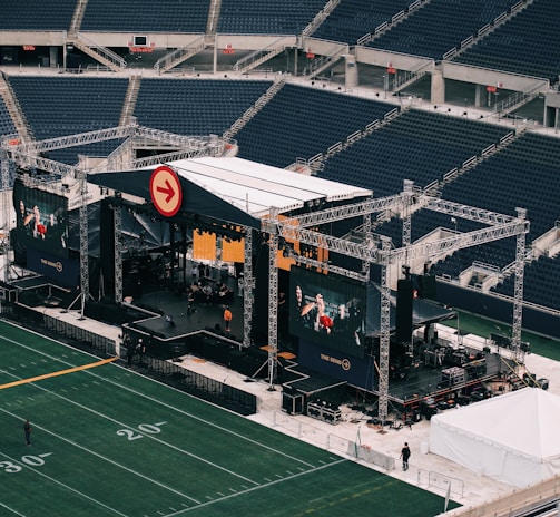 A large outdoor stage is set up inside a stadium, equipped with tall scaffolding, lights, and multiple large display screens. The seats in the stadium are mostly empty. The field is marked with numbers and lines typical of a sports venue. A few people are visible near the stage and on the field.