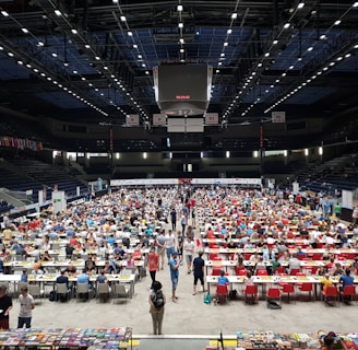 An indoor sports arena filled with rows of tables, each occupied by people engaged in competitive activities. The ceiling is high, with numerous lights illuminating the large space. The audience seating on either side is empty, and there are flags hanging from the upper level.