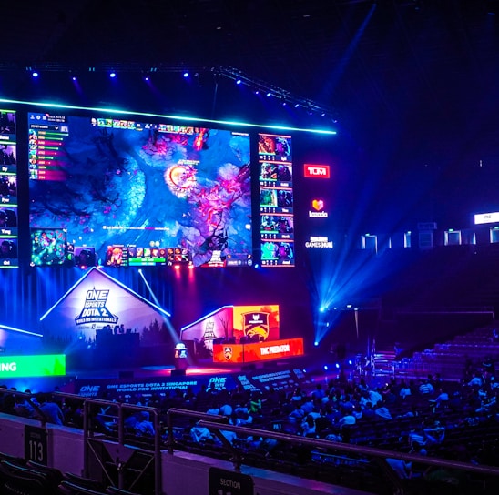 A large esports arena featuring a massive screen displaying a video game, surrounded by sponsor logos and team names. The auditorium is filled with spectators watching the gaming event. Neon blue and purple lights illuminate the stage, creating a vibrant atmosphere.