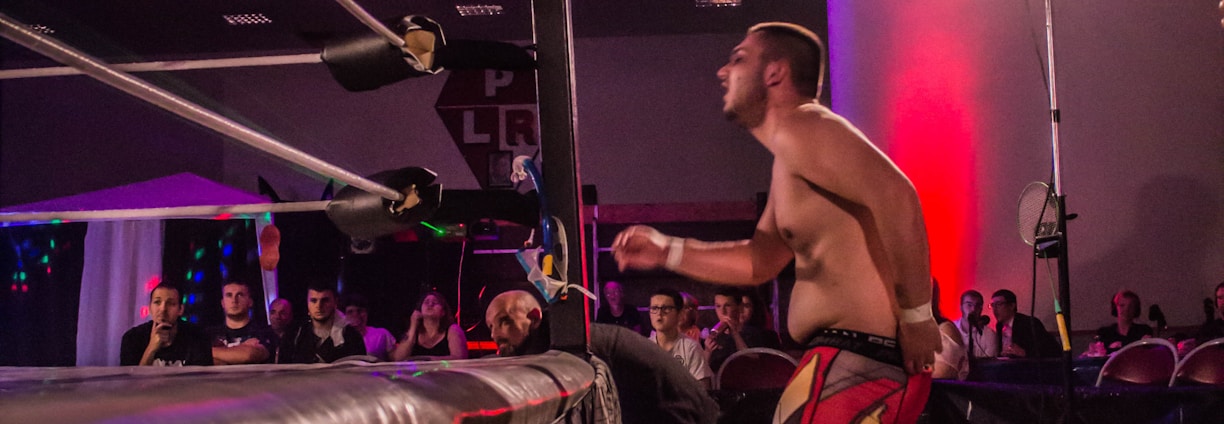 A wrestling match taking place in an indoor venue, where one participant is crouched on the top rope of the ring, while the other stands outside the ring looking at them. A crowd watches the event enthusiastically from their seats.