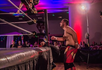 A wrestling match taking place in an indoor venue, where one participant is crouched on the top rope of the ring, while the other stands outside the ring looking at them. A crowd watches the event enthusiastically from their seats.