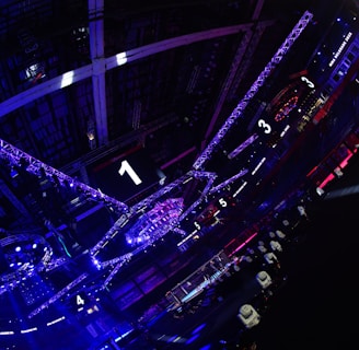 A large indoor arena with colorful and vibrant lighting, featuring a center stage surrounded by rigging and structures. Large numbered screens are visible, indicating marked sections from one to five. Various pieces of equipment and people are scattered around the venue.