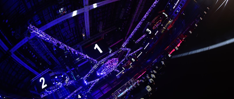 A large indoor arena with colorful and vibrant lighting, featuring a center stage surrounded by rigging and structures. Large numbered screens are visible, indicating marked sections from one to five. Various pieces of equipment and people are scattered around the venue.