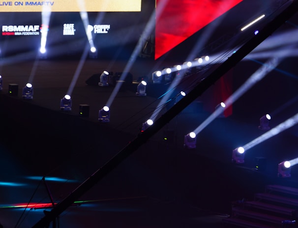 An array of bright stage lights beams across a dark arena, creating a dynamic atmosphere with a large screen displaying various logos and text. A camera crane is visible in the foreground, suggesting a professional event setup.