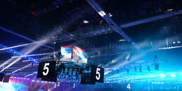 A large indoor arena is equipped with advanced lighting and audio systems. Suspended above the space are numerous digital displays showcasing numbers. The environment is filled with blue illumination and spotlights creating a vibrant atmosphere.