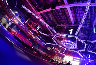 An indoor arena with vibrant purple and blue lighting, featuring large numbered screens suspended from the ceiling. The venue has a high ceiling with intricate metal truss structures and hanging lights, creating a futuristic and energetic atmosphere.
