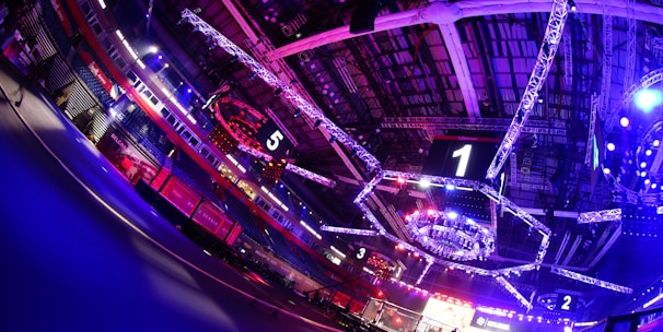 An indoor arena with vibrant purple and blue lighting, featuring large numbered screens suspended from the ceiling. The venue has a high ceiling with intricate metal truss structures and hanging lights, creating a futuristic and energetic atmosphere.