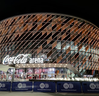 A large, modern arena with a distinctive rounded design featuring a complex pattern of illuminated panels. The facade displays the 'Coca-Cola arena' signage prominently. The venue is surrounded by barriers with 'World Tennis League' branding. The lighting creates a vibrant and inviting atmosphere at night, with lighting around the entrance and inside the building.