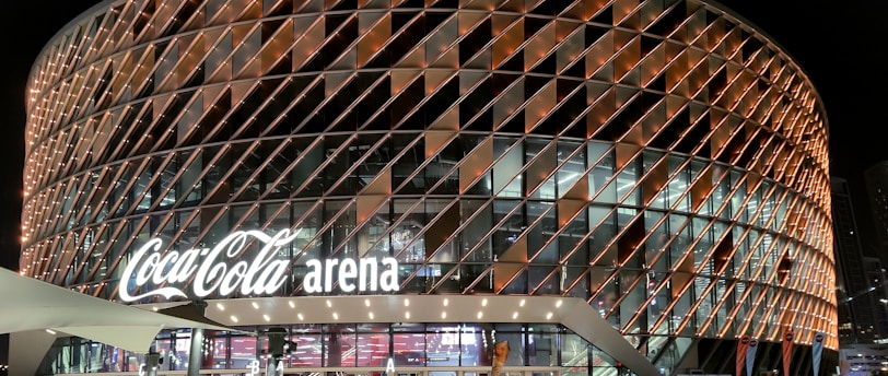 A large, modern arena with a distinctive rounded design featuring a complex pattern of illuminated panels. The facade displays the 'Coca-Cola arena' signage prominently. The venue is surrounded by barriers with 'World Tennis League' branding. The lighting creates a vibrant and inviting atmosphere at night, with lighting around the entrance and inside the building.