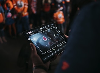 A person is holding a tablet that displays a video or image of a red and blue emblem on the screen. In the background, several people wearing dark and orange colored clothing, possibly sports jerseys, are partially visible. The scene is dimly lit, suggesting an indoor or evening setting.
