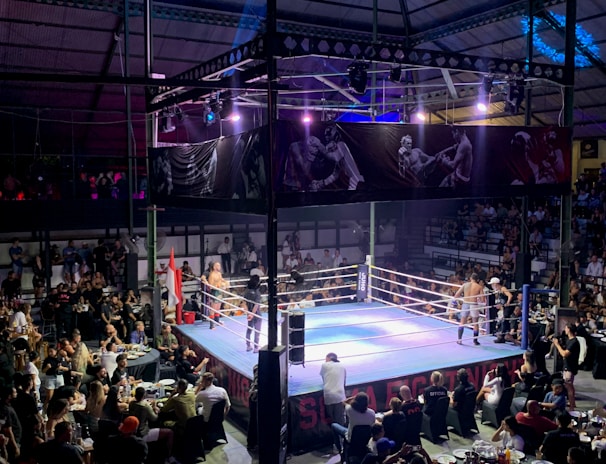 A crowded indoor arena with a boxing ring at the center, surrounded by an audience seated at tables. The scene is illuminated by overhead lights, casting a vibrant glow over the ring where two fighters are preparing for a match. Spectators are engaged, capturing the event with cameras and enjoying the lively atmosphere.