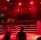 A wrestling ring is illuminated with red lighting, creating a dramatic atmosphere. Several people are visible around the ring, with silhouetted spectators in the foreground. Banners or art adorn the upper walls, and the venue has an industrial ceiling with exposed beams.