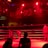 A wrestling ring is illuminated with red lighting, creating a dramatic atmosphere. Several people are visible around the ring, with silhouetted spectators in the foreground. Banners or art adorn the upper walls, and the venue has an industrial ceiling with exposed beams.