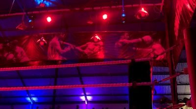 A brightly lit boxing or wrestling ring with vivid red and blue lights. Banners with images of fighters hang from the ceiling. The ropes of the ring are prominently visible, and a stool is placed inside the ring. The arena is filled with an audience, silhouetted against the colorful lighting.