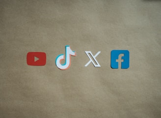 Four social media platform logos are arranged on a brown background. From left to right, the logos are for YouTube in red, TikTok in a combination of red, blue, and white, X in white, and Facebook in blue.
