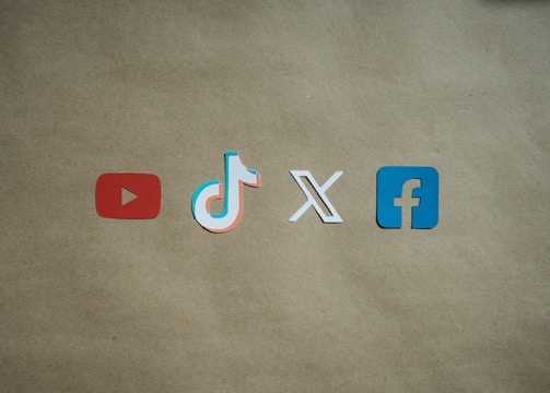 Four social media platform logos are arranged on a brown background. From left to right, the logos are for YouTube in red, TikTok in a combination of red, blue, and white, X in white, and Facebook in blue.