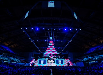 An expansive venue filled with a large audience, featuring a grand stage lit with vibrant blue and pink lights. The ceiling is decorated with a large display, and numerous screens showcase the event's branding. The atmosphere is dynamic and futuristic, reflecting a technologically advanced conference.
