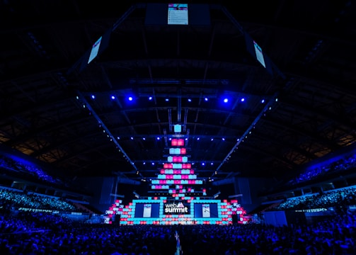 An expansive venue filled with a large audience, featuring a grand stage lit with vibrant blue and pink lights. The ceiling is decorated with a large display, and numerous screens showcase the event's branding. The atmosphere is dynamic and futuristic, reflecting a technologically advanced conference.