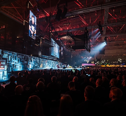 A large indoor event with a stage featuring bright screens and spotlights, attended by a substantial crowd. The venue has a high ceiling with industrial beams and red lighting accents. The atmosphere resembles a concert or a conference with a focus on visual effects.