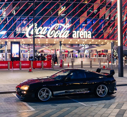 A sleek black sports car is parked in front of an illuminated arena entrance, with a large sign bearing the name of a famous beverage brand. The building features a modern, geometric facade with bright red and blue lights, creating a vibrant and lively atmosphere.